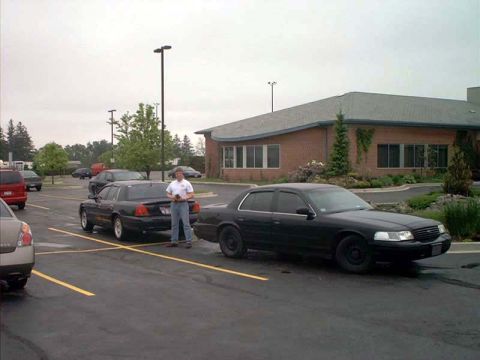 2000 Ford Crown Victoria - Two black Ford Crown Victorias are parked in a lot near a brick building. A person stands between them.