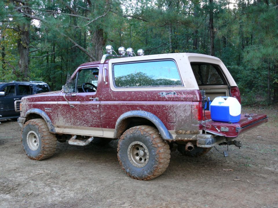 Muddy maroon 1988 Ford Bronco with off-road tires, tailgate down, cooler in back. Another dark SUV partially visible