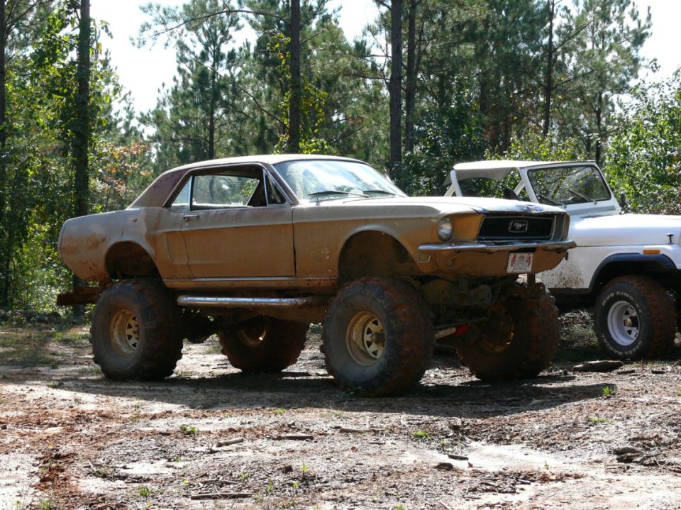 Mud-covered 1967 Ford Mustang coupe lifted on large off-road tires, parked next to a white Jeep