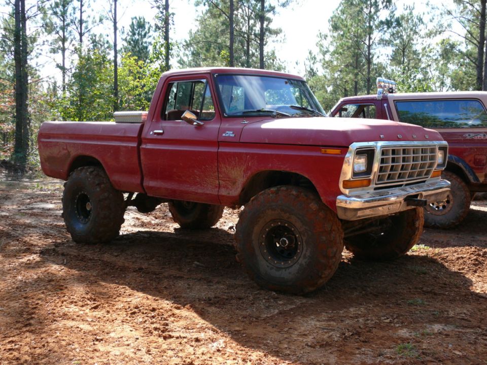 Red pickup truck with large off-road tires parked on dirt, another vehicle visible behind it