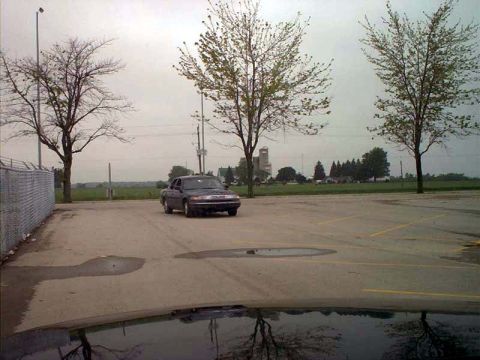 2000 Ford Crown Victoria - A dark sedan is parked in an empty lot with trees and buildings visible in the distance.