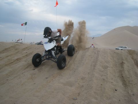 1996 Ford Bronco - Rider on an ATV navigates a sand dune, creating a dust cloud. A pickup truck is visible in the distance.