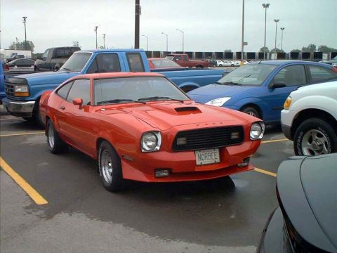 2000 Ford Crown Victoria - This orange 1975 Ford Mustang II coupe is parked in a lot with other vehicles, including a blue Ford Focus and a blue pickup truck.