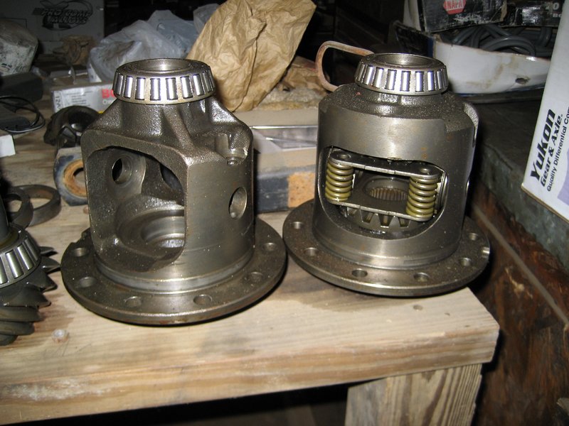 Two differential carriers and gears sit on a wooden surface. One carrier is open, showing internal gears. The other carrier has springs visible