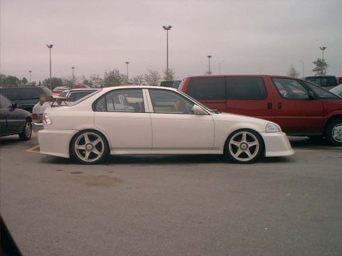 2000 Ford Crown Victoria - A white sedan with aftermarket wheels and a body kit is parked in a lot under an overcast sky.