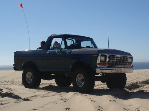 1979 Ford Bronco - A dark blue Ford Bronco with large tires drives on sand dunes under a clear blue sky.