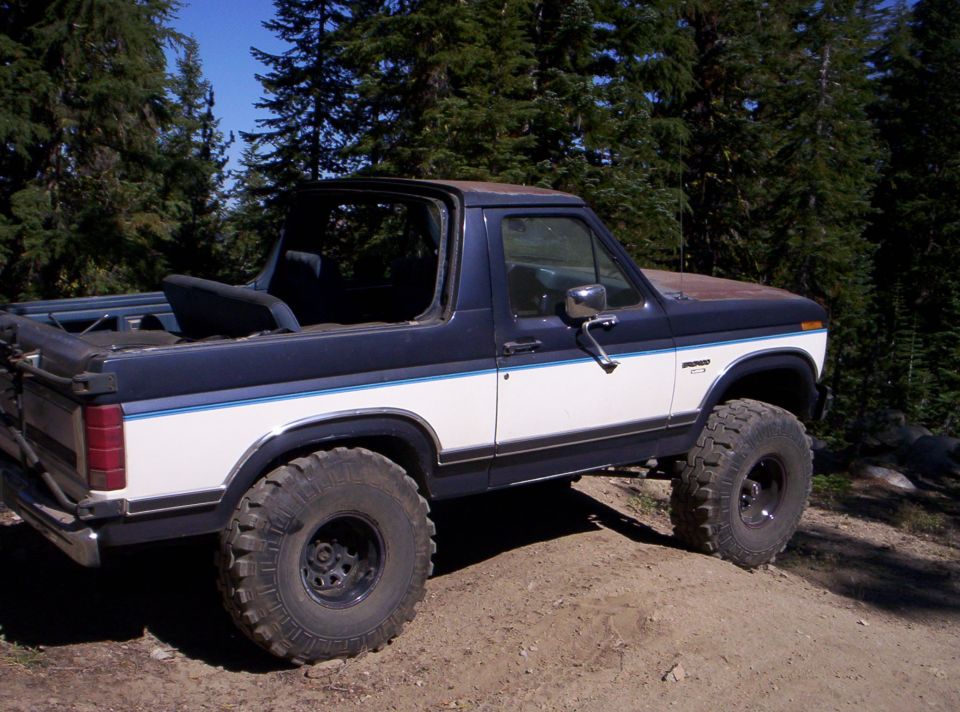 Lifted 1985 Ford Bronco with aftermarket wheels and tires, parked on a dirt road with trees in the background