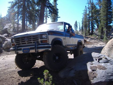1985 Ford Bronco - A lifted Ford Bronco navigates a rocky trail under a clear blue sky.