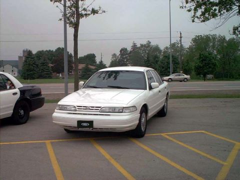 2000 Ford Crown Victoria - A white 2000 Ford Crown Victoria is parked in a lot, with a black and white police car to its left.