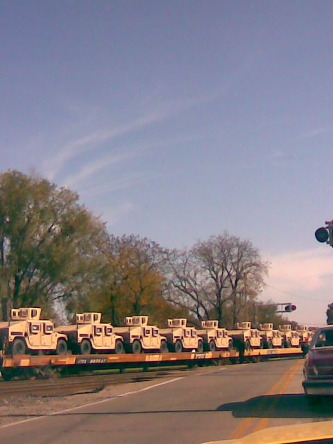 1994 Ford Bronco - Multiple tan military trucks are transported on flatbed train cars under a partly cloudy sky.