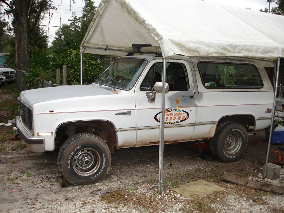 White GMC K5 Blazer with a canopy, parked under a tent. It has a 'Putnam County Speedway' decal on the door
