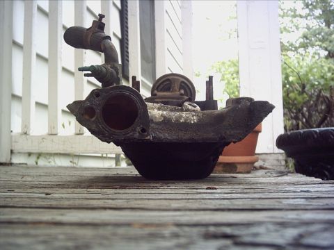1979 Ford LTD - A weathered engine intake manifold rests on a wooden deck, with a house and greenery in the background.