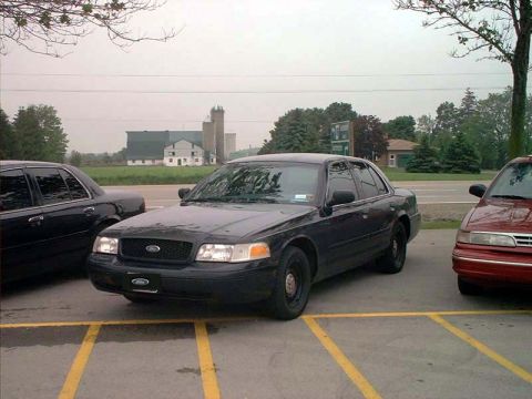 2000 Ford Crown Victoria - Two sedans parked in a lot, one black Ford Crown Victoria and a red car, with rural buildings visible behind them.