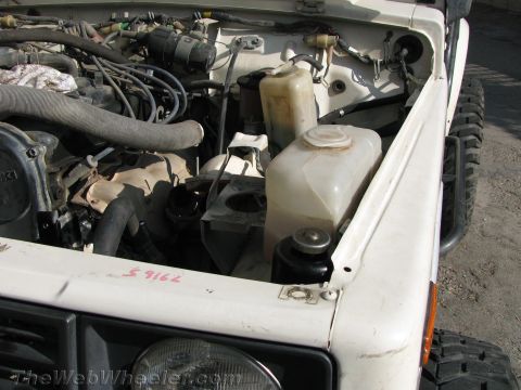 1987 Suzuki Samurai - The engine bay of a white 1987 Suzuki Samurai, showing various components and fluid reservoirs.