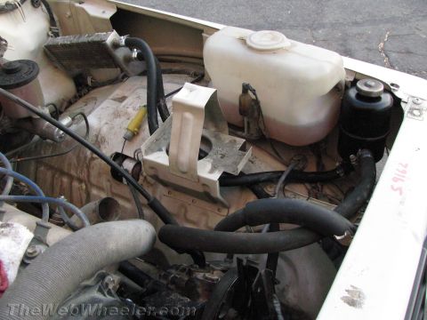 1987 Suzuki Samurai - Engine bay view of a Suzuki Samurai showing a white plastic coolant/washer reservoir, a black cylindrical reservoir at right, various black…