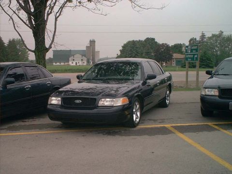 2000 Ford Crown Victoria - Two sedans are parked in a lot with buildings and trees in the background under an overcast sky.