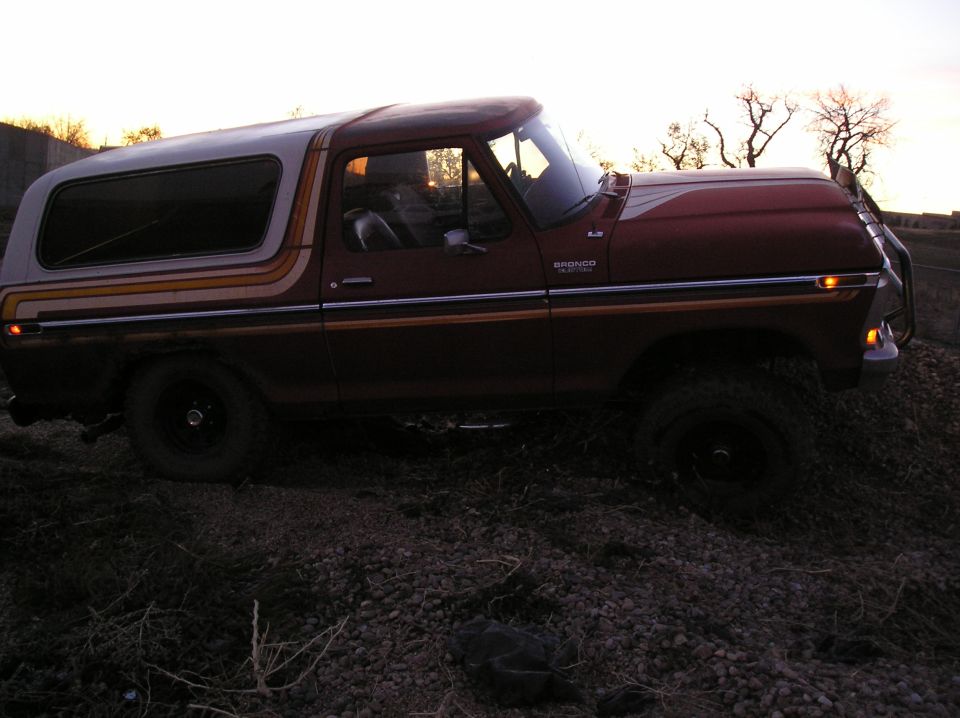 Side view of a red Ford Bronco with a white top and striped decals. It's parked on gravelly ground