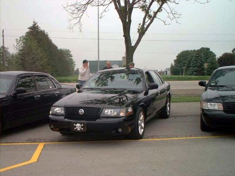 2000 Ford Crown Victoria - Two black sedans are parked in a lot under a tree, with two men standing near the front of the car on the right.
