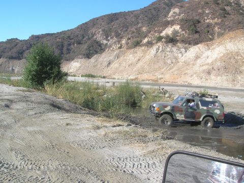 1985 Ford Bronco - A camouflage Ford Bronco navigates a muddy terrain with a mountainous backdrop.
