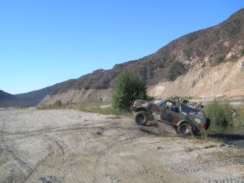 1985 Ford Bronco - This camouflage Ford Bronco navigates a dusty, off-road terrain with a mountainous backdrop. Text is visible on the vehicle's door.