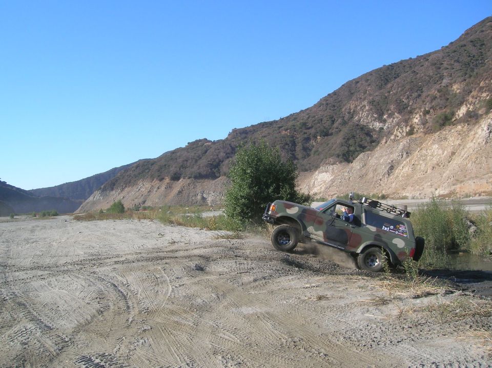 Camouflage Ford Bronco driving off-road, 3q-front view. Visible text on door