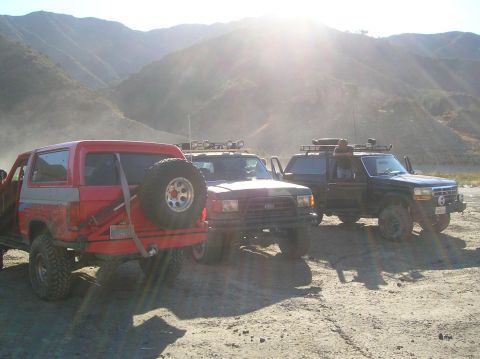 1985 Ford Bronco - Three Broncos are parked on a dusty road with mountains in the background.