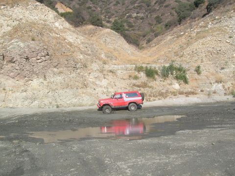 1985 Ford Bronco - A red SUV with a spare tire on the back is parked on a dirt road with a puddle reflecting its image.