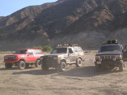 1985 Ford Bronco - Three Ford Broncos are parked on a dusty, arid terrain with mountains in the background. The vehicles appear to be from the late 1980s or e…