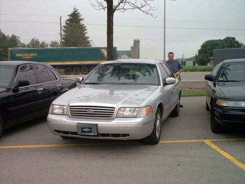 2000 Ford Crown Victoria - A silver 2000 Ford Crown Victoria is parked next to a black car, with a man standing by the driver's side.