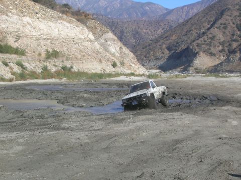1985 Ford Bronco - A white pickup truck is stuck in a muddy riverbed with mountains in the background.