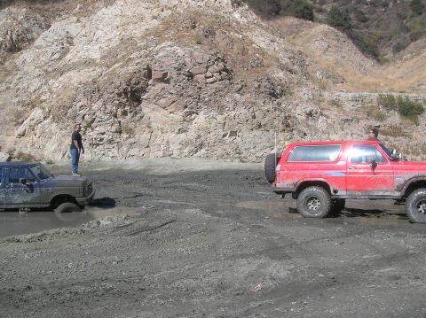 1985 Ford Bronco - Two SUVs, including a red 1985 Ford Bronco, are stuck in a muddy area with a rocky hillside.