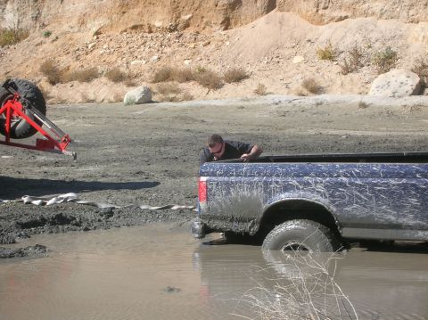 1985 Ford Bronco - A person attempts to free a stuck pickup truck from a muddy water hole while another vehicle waits.