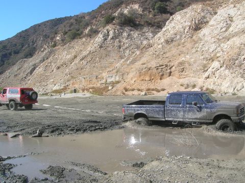 1985 Ford Bronco - Two off-road vehicles are stuck in a muddy area with a rocky hillside in the background.