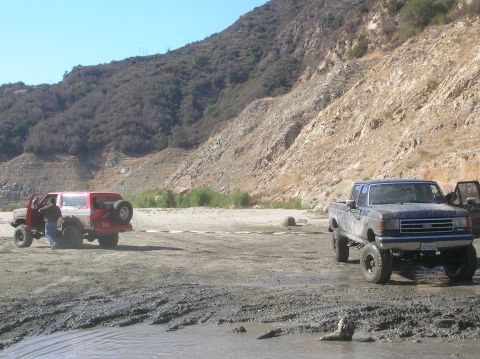 1985 Ford Bronco - Two off-road vehicles are parked in a muddy, rocky area with a large hillside in the background.