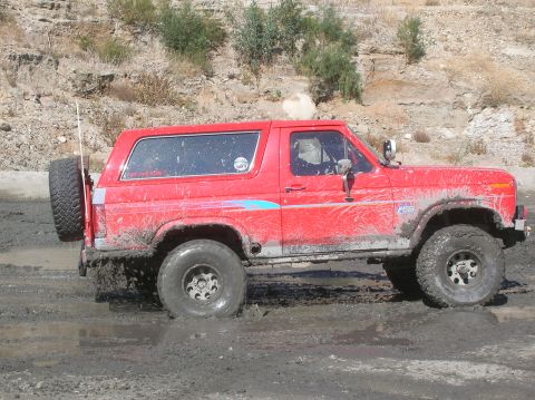 1985 Ford Bronco - Red Ford Bronco navigating through a muddy terrain, with its tires deep in the mire.