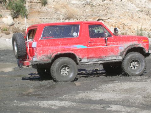 1985 Ford Bronco - This red SUV is shown off-road, navigating through a muddy terrain with its tires caked in dirt.