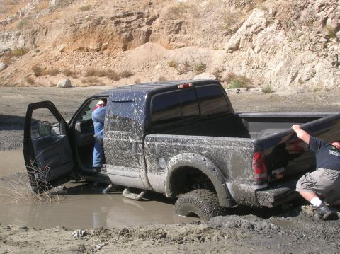 1985 Ford Bronco - A black pickup truck is stuck in a muddy area, with one person exiting and another attempting to push.
