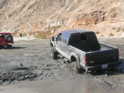 1985 Ford Bronco - A pickup truck is stuck in thick mud, with a red Jeep in the background on a dirt road.