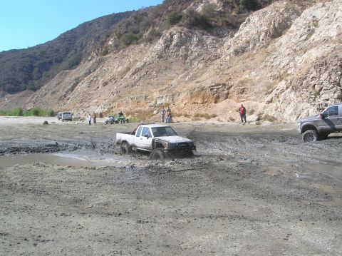 1985 Ford Bronco - Multiple vehicles navigate a muddy off-road course with steep hills in the background.