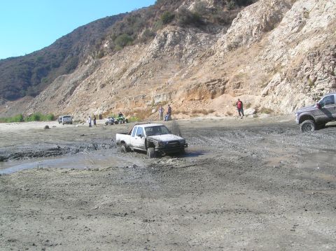 1985 Ford Bronco - Several off-road vehicles are stuck in a muddy area with a rocky hillside in the background.