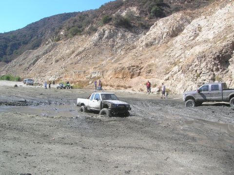 1985 Ford Bronco - Several vehicles are stuck in a muddy terrain with hills in the background.