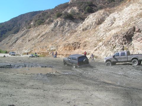 1985 Ford Bronco - A dark SUV struggles through a muddy off-road course, with a large pickup truck visible in the background.