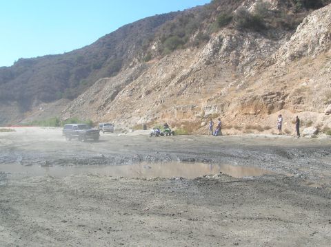 1985 Ford Bronco - Several vehicles and people are gathered in a muddy, off-road area with a large hill in the background.