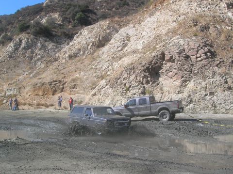 1985 Ford Bronco - Two off-road vehicles navigate a muddy terrain with spectators observing from the side of a rocky hill.