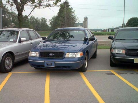 2000 Ford Crown Victoria - Blue Ford Crown Victoria with Ford grille badge parked in striped lot stalls between silver and dark sedans, rural backdrop.