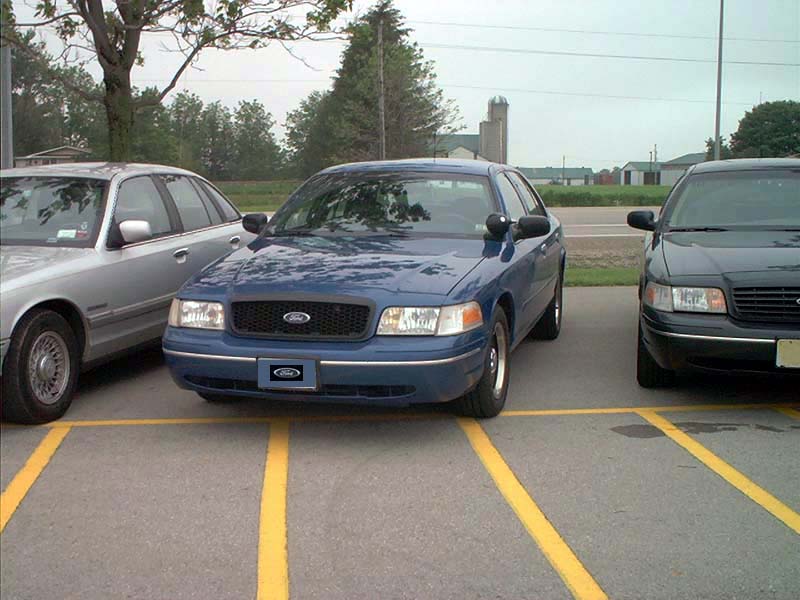 Blue Ford Crown Victoria parked in a lot between two other Crown Victorias
