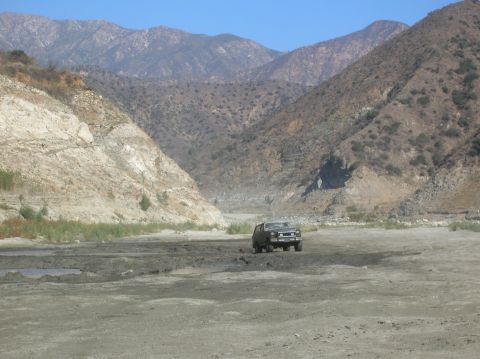 1985 Ford Bronco - A dark SUV navigates a dry riverbed in a mountainous canyon under a clear blue sky.