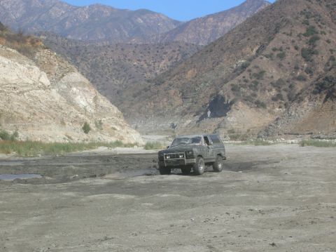 1985 Ford Bronco - Ford Bronco driving through a dry riverbed with mountainous terrain.