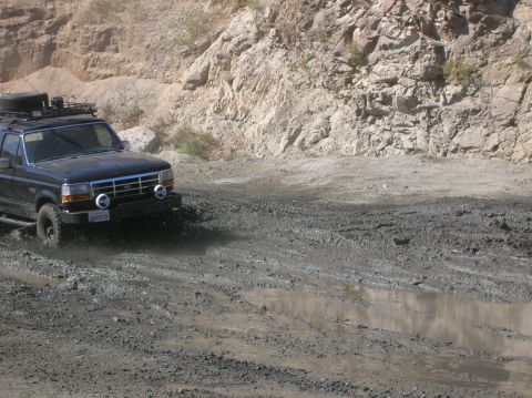 1985 Ford Bronco - This Ford Bronco navigates a muddy off-road trail with rocky hills in the background.