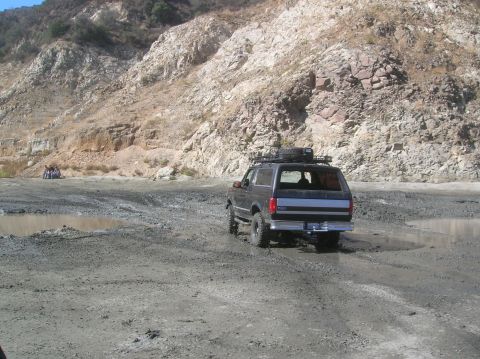 1985 Ford Bronco - A Ford Bronco navigates a muddy, rocky terrain with a steep, dry hillside in the background.